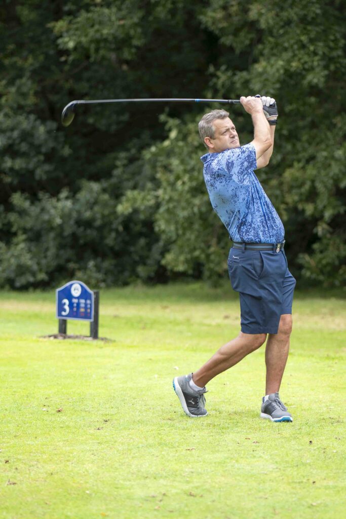Client Event Day with Cullen Wealth and Cheadle Sports Club at Bramhall Park Golf Course. The image features a gentleman winging his club through the air as he strikes the golf ball of the tee. he finishes his motion over his head. The individual is wearing a blue, floral top with similar coloured shorts.