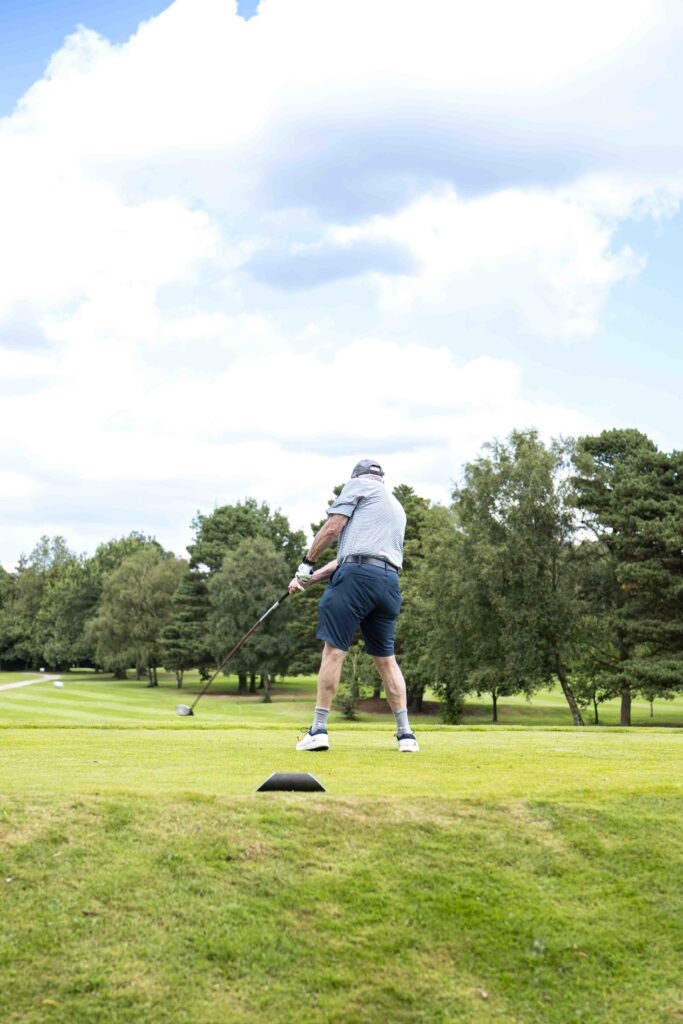 Client Event Day with Cullen Wealth and Cheadle Sports Club at Bramhall Park Golf Course. The image features a gentle swinging his club towards the golf ball, situated on the tee. He is wearing a white shirt with a blue pair of shorts.